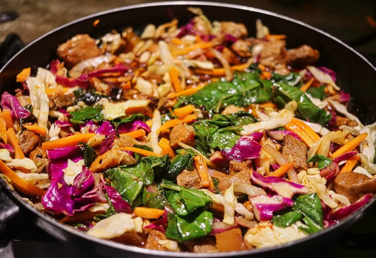 Colorful homemade stir fry with chicken, carrots, spinach, cabbage, and purple cabbage cooking in a skillet. A healthy, budget-friendly meal idea for beginners.