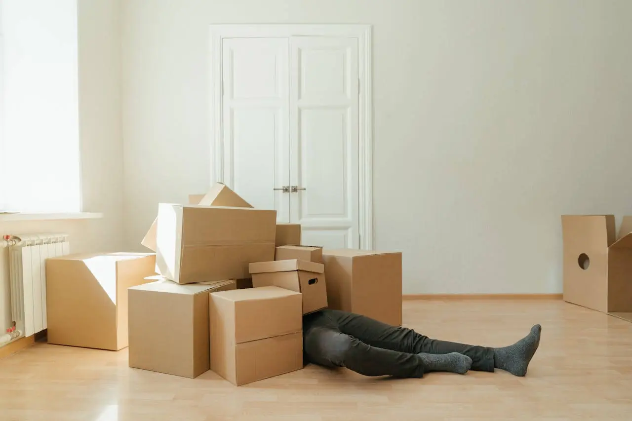 Person lying on the floor covered in moving boxes, symbolizing the stress and exhaustion of moving into a first apartment.